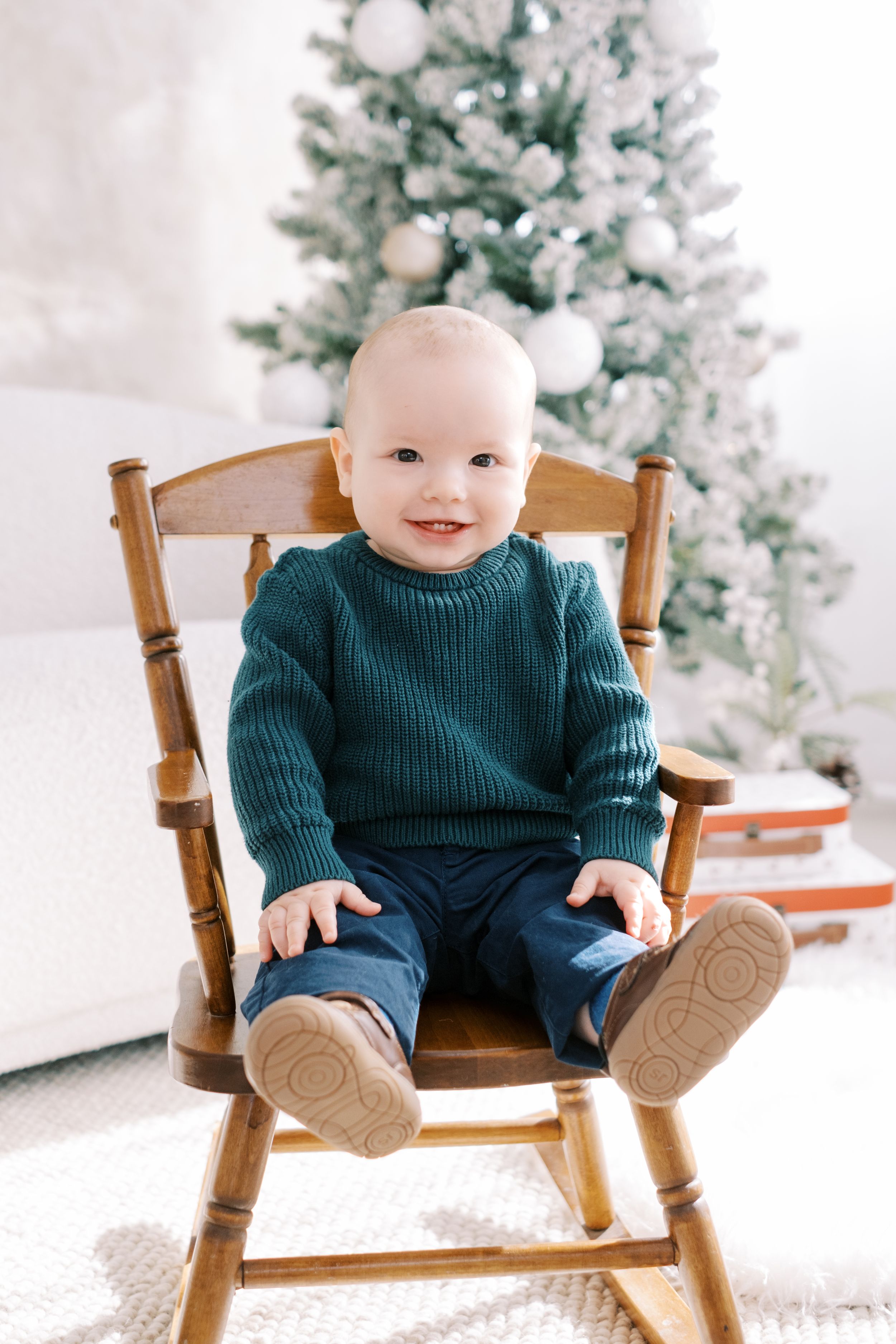 Small child sits in wooden rocking chair wearing navy sweater and pants with white winter decor in background.