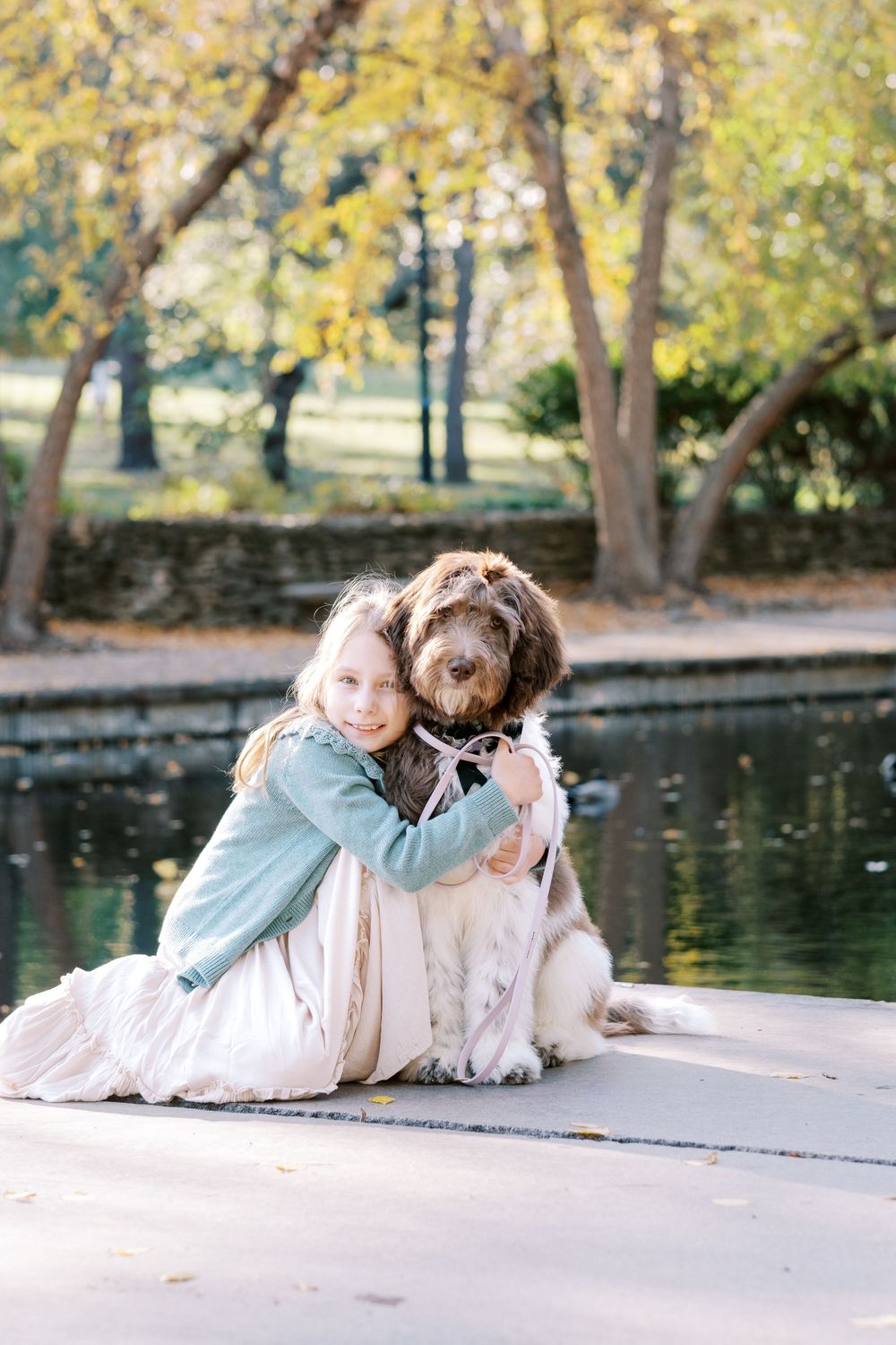 A person and a dog relax together by a tranquil pond surrounded by autumn trees with golden foliage.