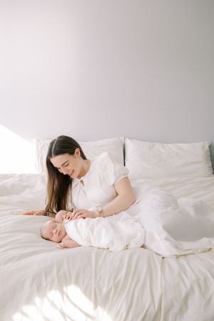 A tender moment captured in soft white tones as a mother lies on a bed with her newborn baby in matching light attire.