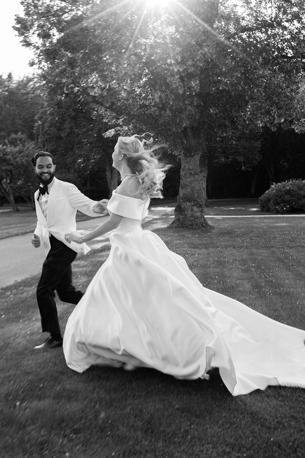A couple in wedding attire playfully spins and dances together on a grassy lawn during golden hour sunlight.