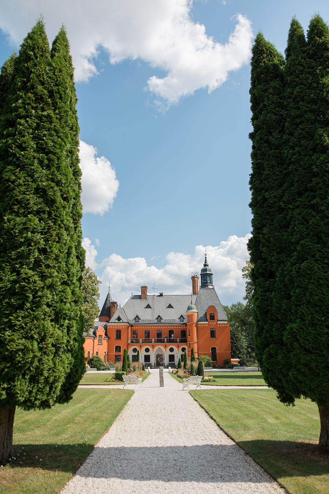 A grand historic mansion with red brick and grey stone facade viewed through tall cypress trees along a gravel path.