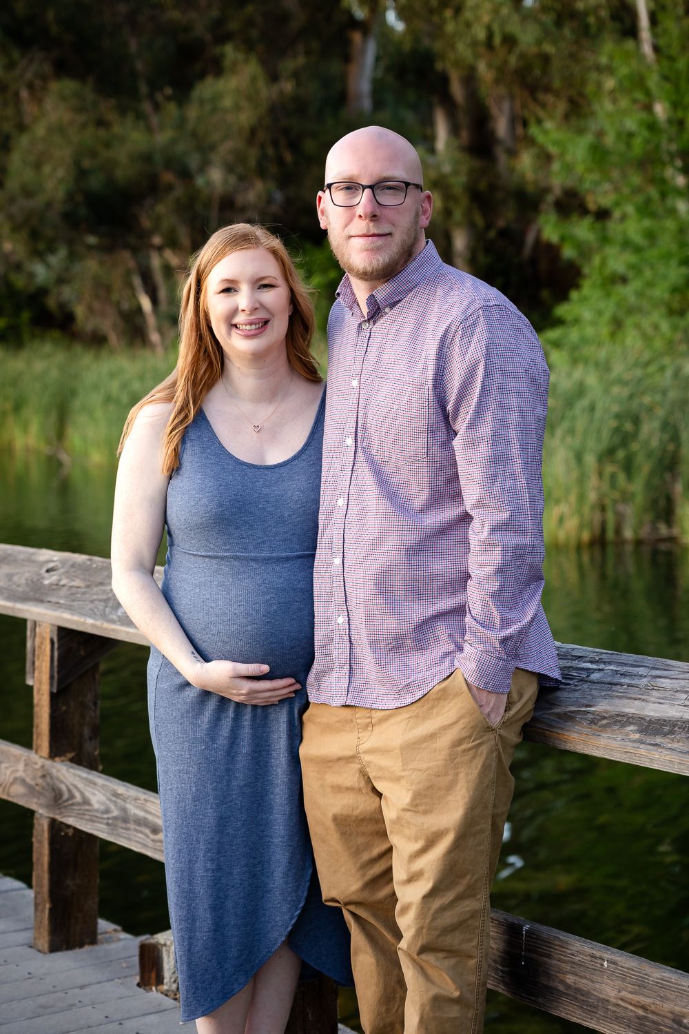 A couple poses together on a wooden dock by a serene pond during a maternity photo session.