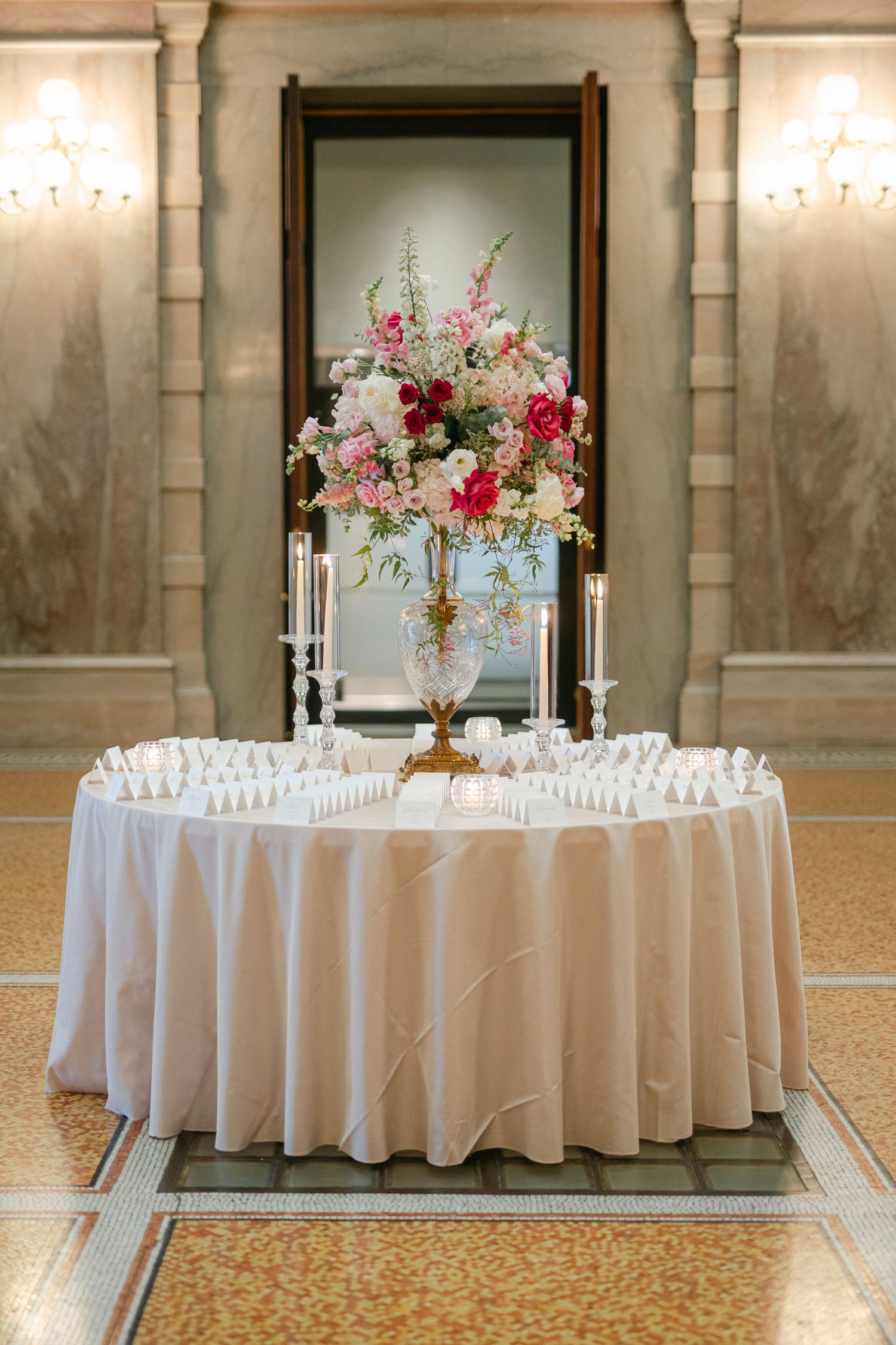 Elegant wedding reception tables with white linens and tall pink floral centerpieces in a grand ballroom with patterned carpet.