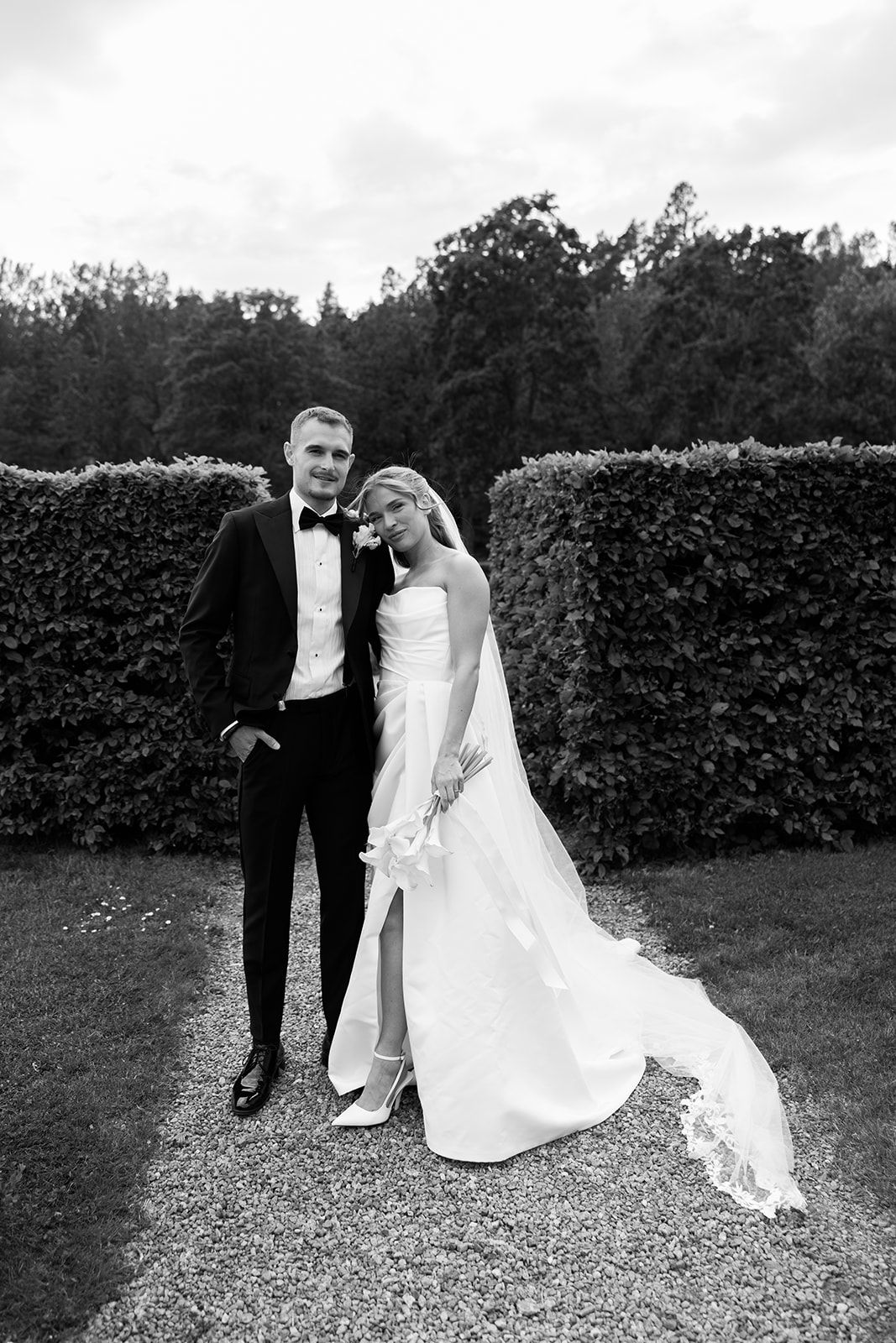 A black and white wedding photo showing a couple posing outdoors against a manicured hedge background.