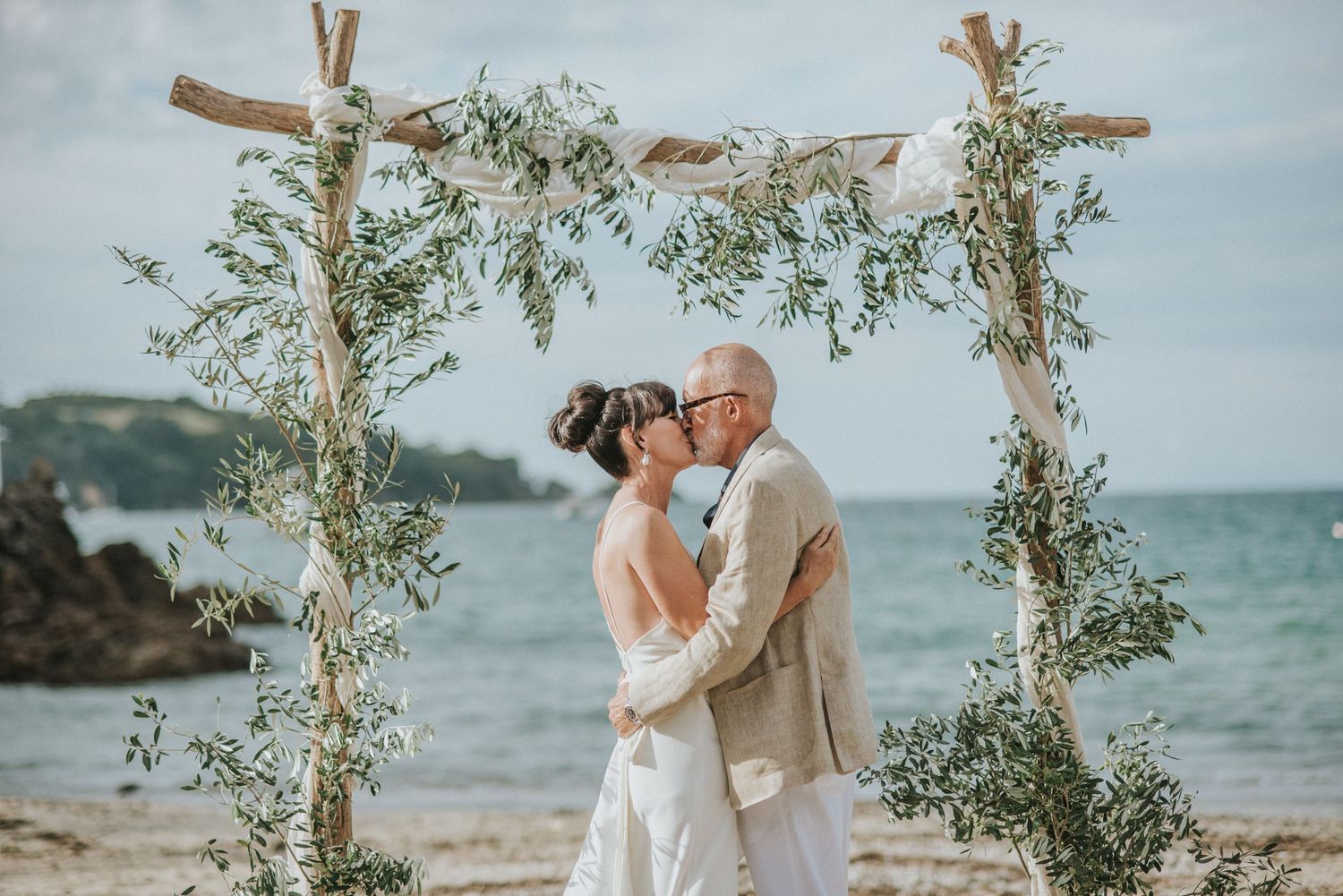 A couple in white and pink attire poses joyfully in a green outdoor setting on a cloudy day.