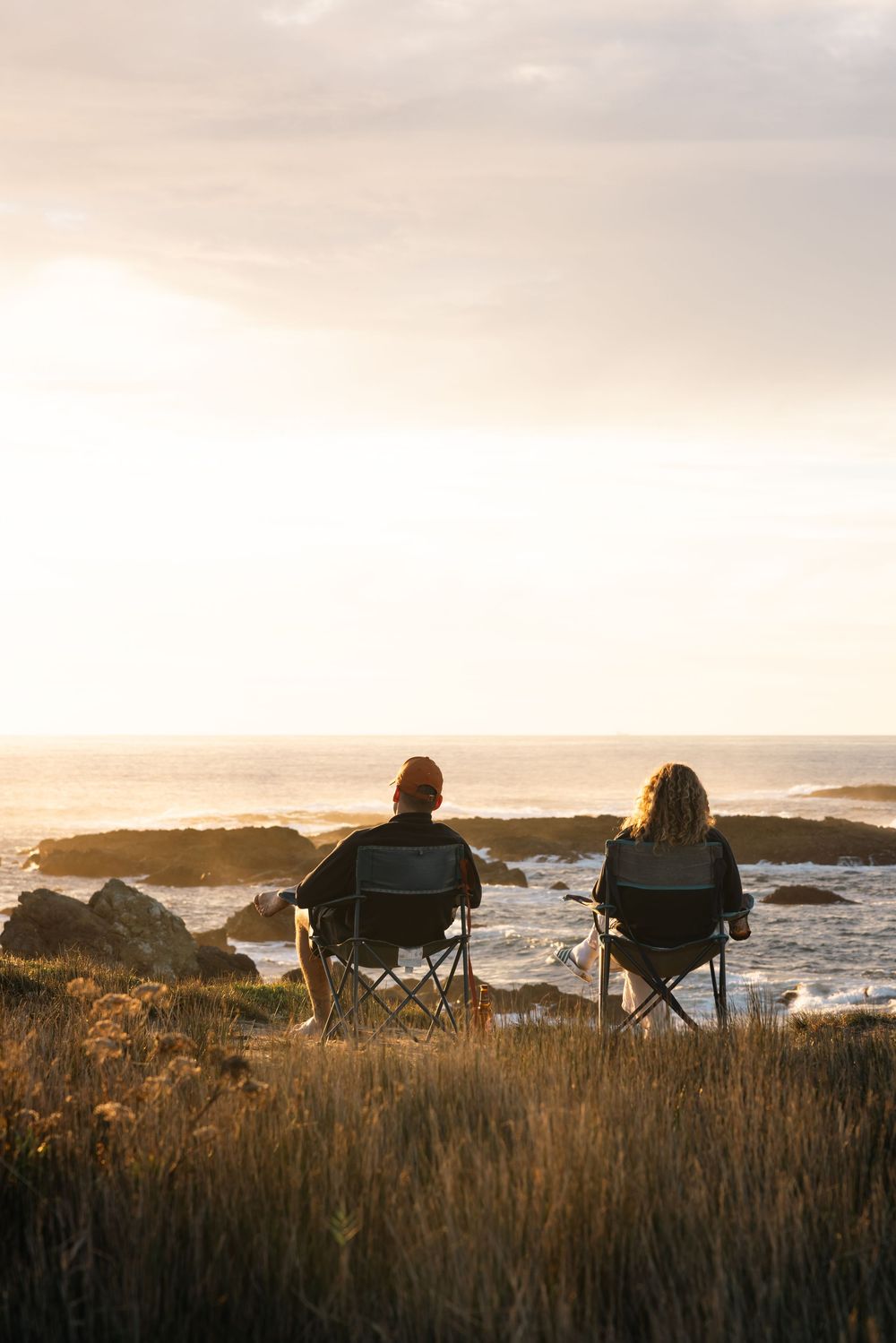 Camping chairs silhouetted against a dramatic sunset over coastal cliffs and ocean waves.