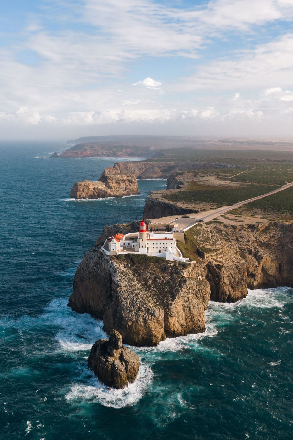 Aerial view of a lighthouse perched on rugged coastal cliffs with crashing waves below.