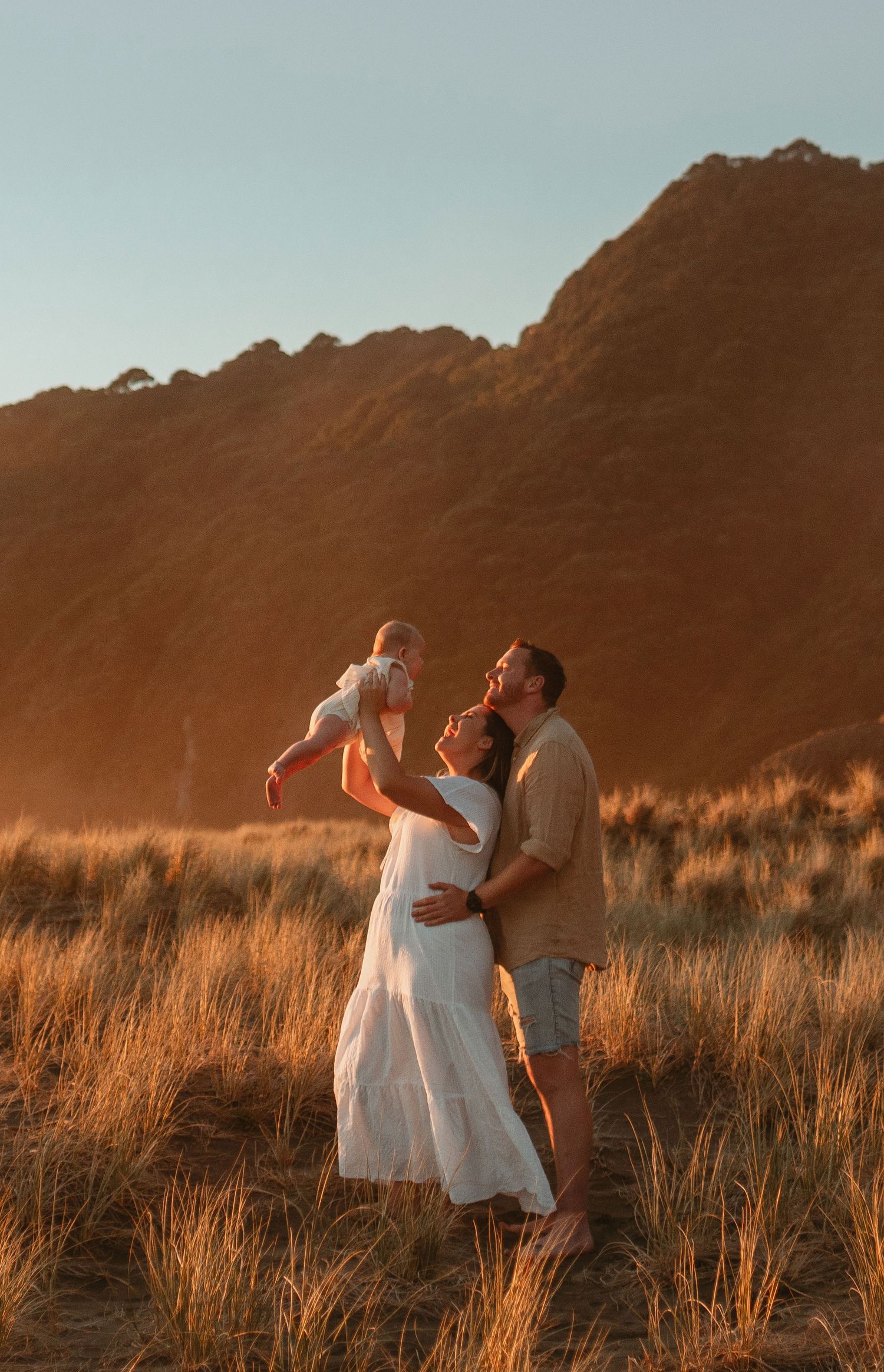 A couple in white dresses embraces in a golden field at sunset with mountains in the background.