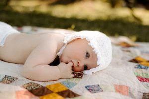 Infant resting on quilted blanket wearing delicate white dress with natural lighting.