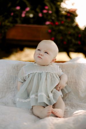 Baby wearing white ruffled dress lying on white linens with garden backdrop.