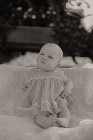 Black and white portrait of baby in white dress lying on soft blanket outdoors.