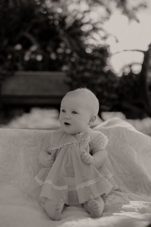 Monochrome image of baby in white dress on textured blanket with bokeh background.