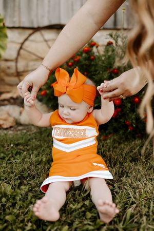 Baby wearing orange cheerleader outfit lying on grass with hands reaching into frame.