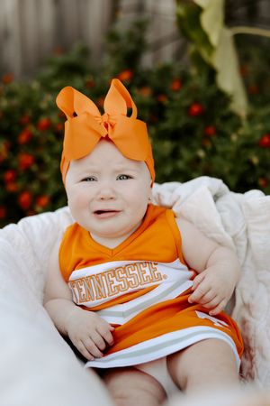 Baby in bright orange cheerleader uniform with matching bow lying on white blanket outdoors.