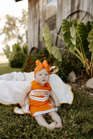 Baby wearing orange Tennessee outfit sits on grass near tropical plants.