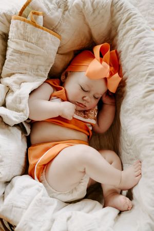 Sleeping baby in orange outfit and bow rests on white blankets.