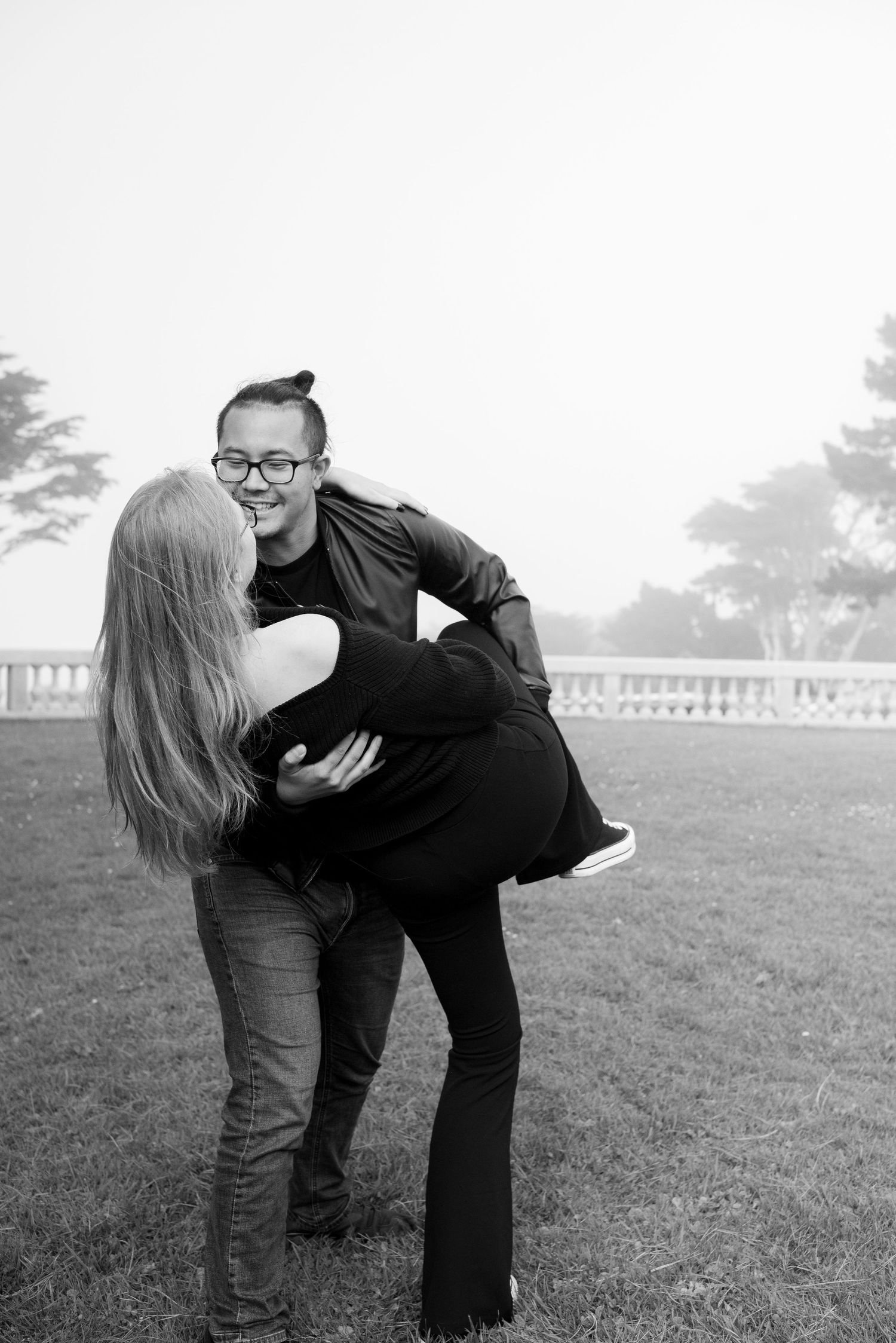 A playful black and white photo of a couple laughing together on a grassy field.