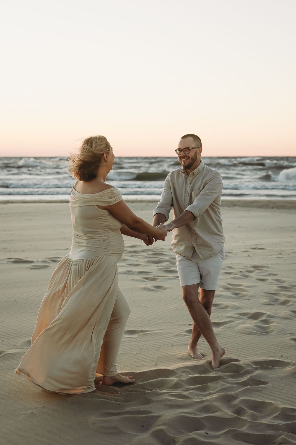 A couple dances together on a sandy beach at sunset with ocean waves in the background.