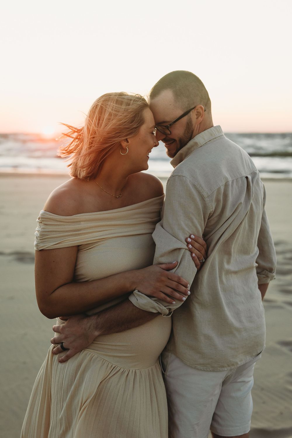 A romantic couple embraces on a beach at sunset wearing light colored outfits.
