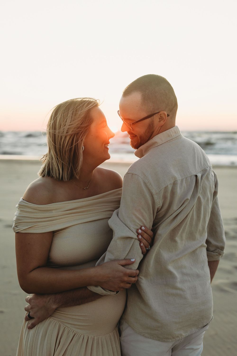 A romantic couple embraces on the beach at sunset, wearing light colored clothing and sharing an intimate moment.
