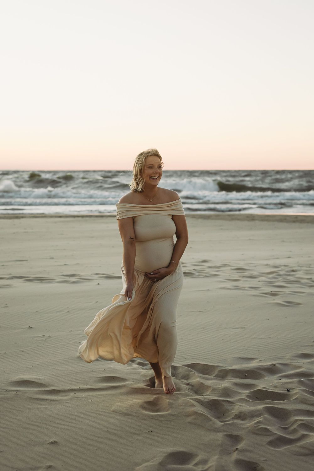 A woman in a flowing beige dress twirls gracefully on a sandy beach at sunset as waves crash in the background.