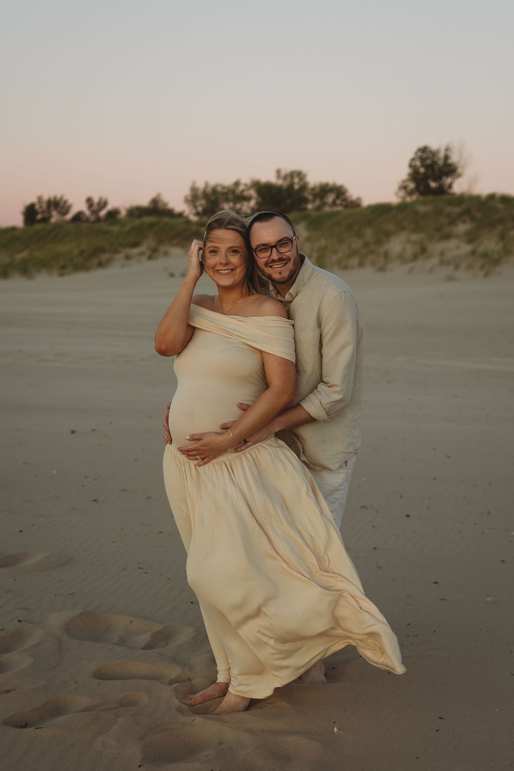 A couple shares tender moments during a beach maternity photoshoot at sunset with sand dunes in the background.