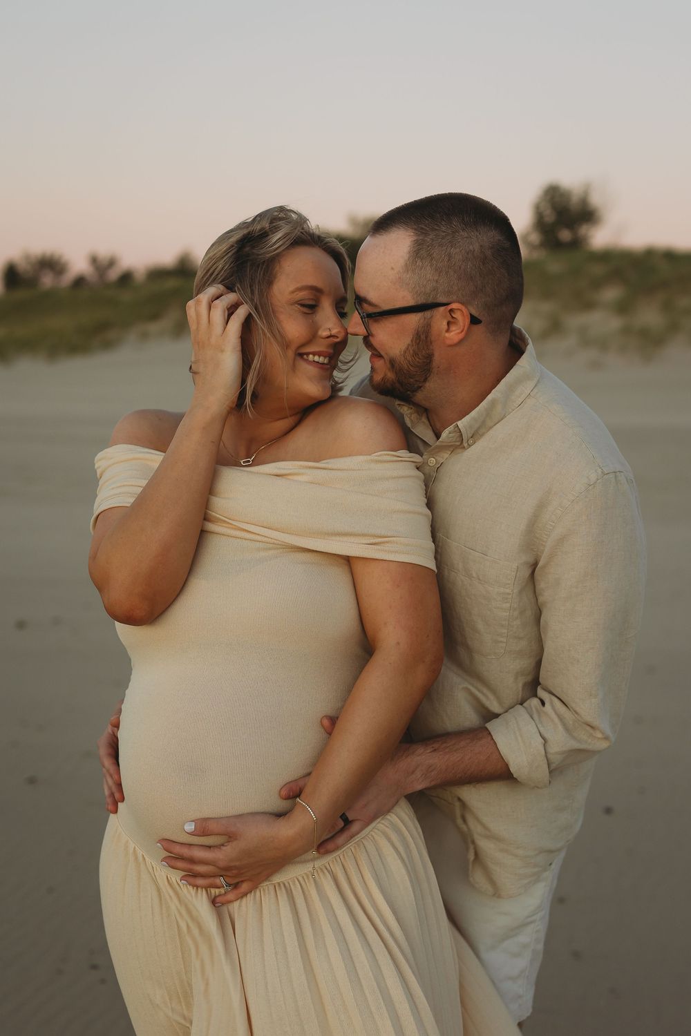 A couple embraces intimately on a sandy beach at sunset wearing light colored clothing.