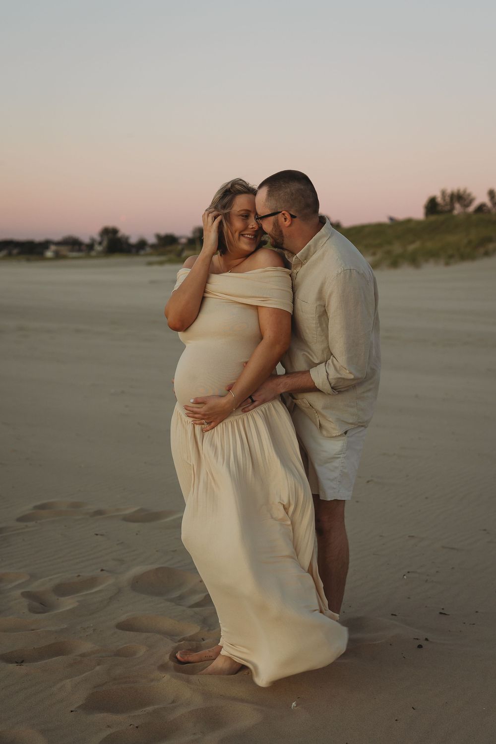 A couple embraces on a sandy beach at sunset, with the pregnant woman wearing a flowing cream-colored dress.