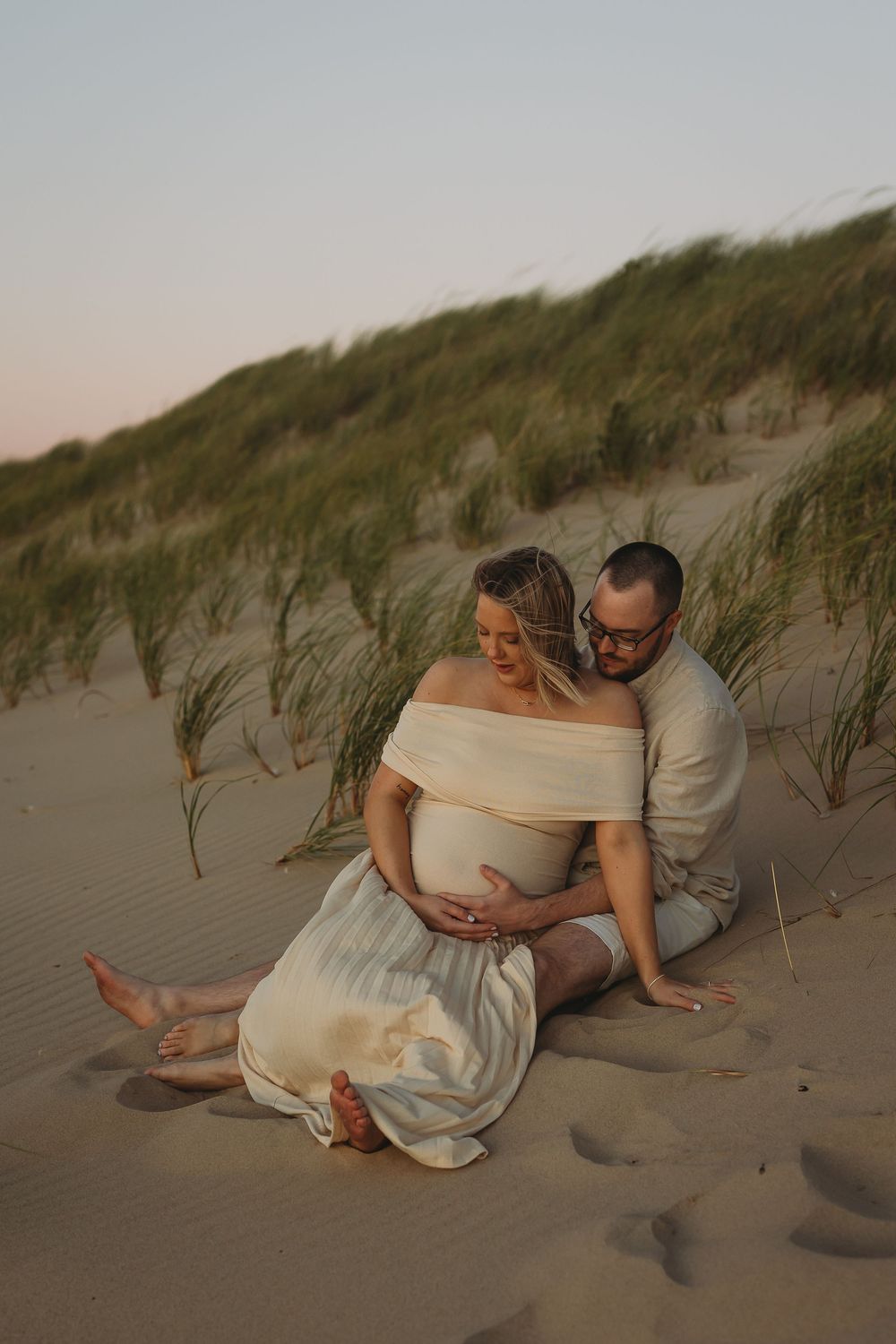 A romantic maternity photo session at sunset on a sandy beach with tall grass dunes in the background.