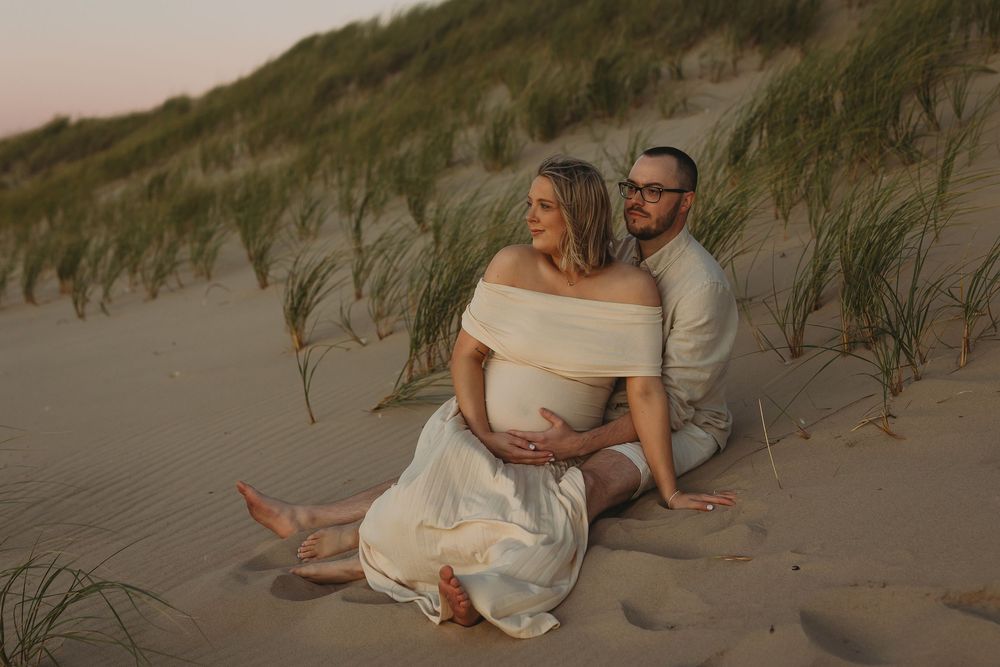 Romantic couple in white outfits sitting together on sandy beach dunes at sunset.