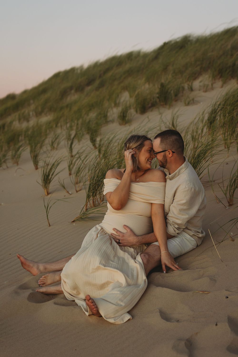 Romantic couple embraces on sandy beach dunes at sunset with tall grass swaying in the background.