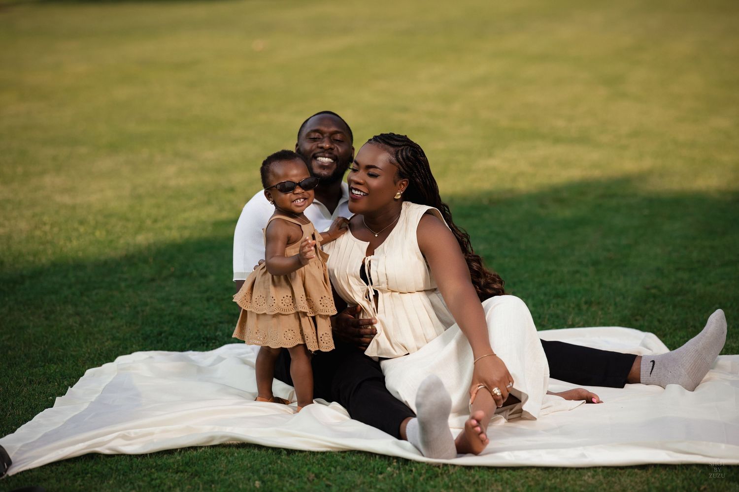 A family sits together on a white blanket in the grass during a sunny outdoor photo session.