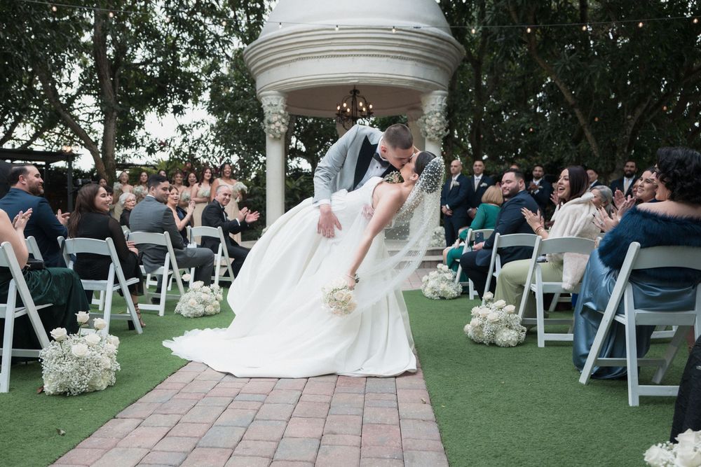 Groom dips bride for a romantic kiss at outdoor wedding ceremony with white chairs and floral decorations.