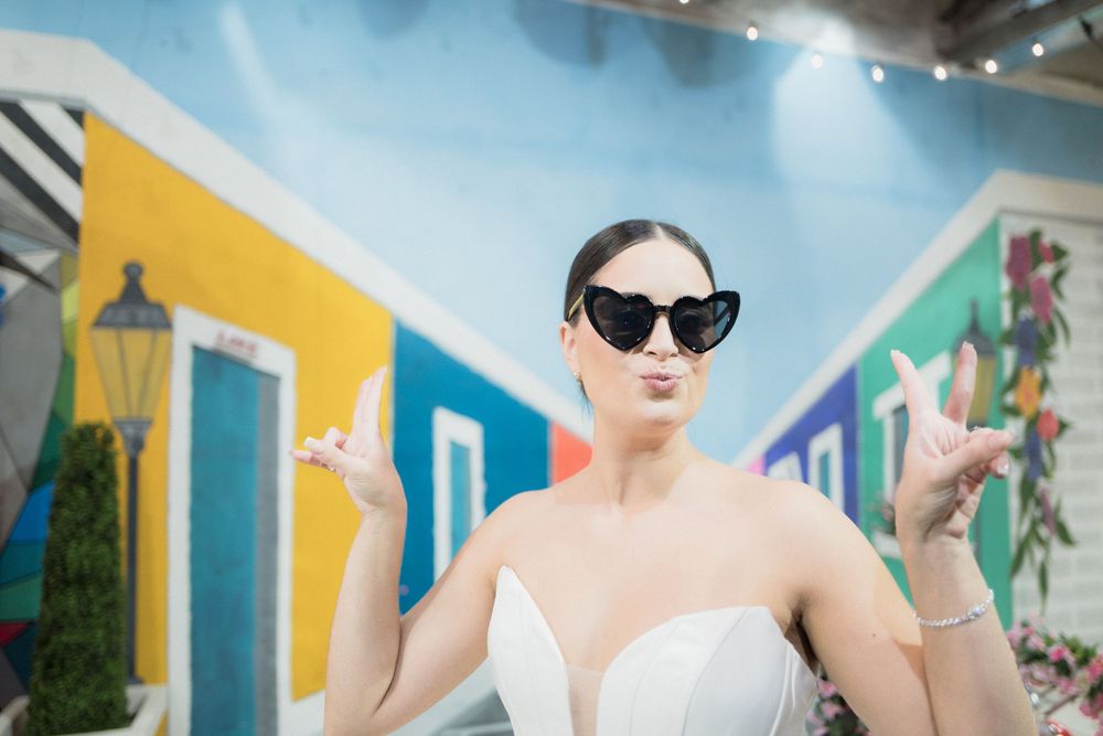 Woman in white dress and cat-eye sunglasses posing playfully in front of colorful tropical mural backdrop.