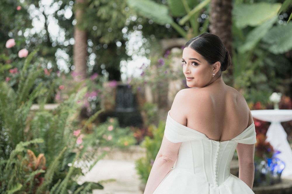 Bride in elegant off-shoulder white wedding gown poses in lush garden setting with tropical greenery.