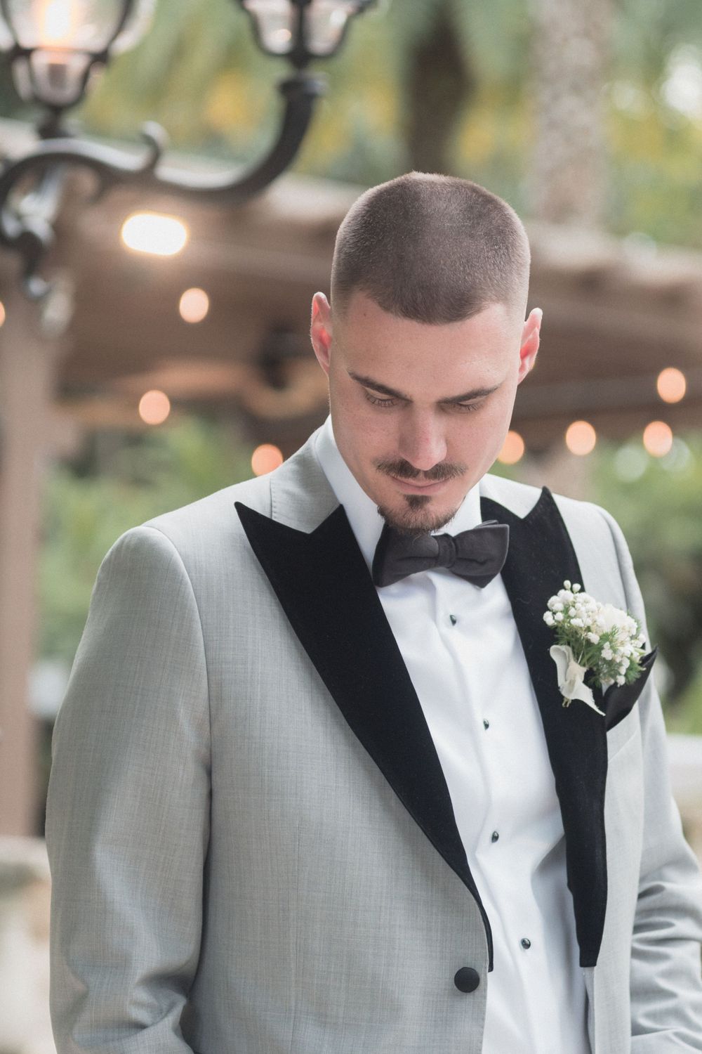 Groom in gray tuxedo with black lapels, bow tie, and floral boutonniere looking down at outdoor wedding venue.