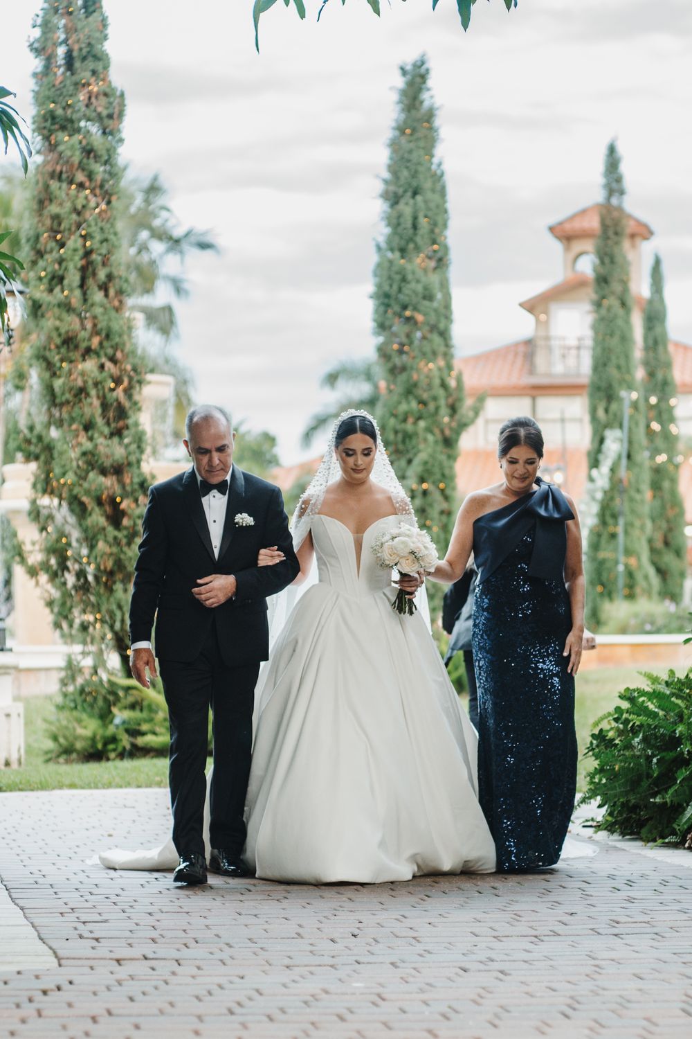 Wedding party walking together down a stone path lined with cypress trees at an elegant venue.