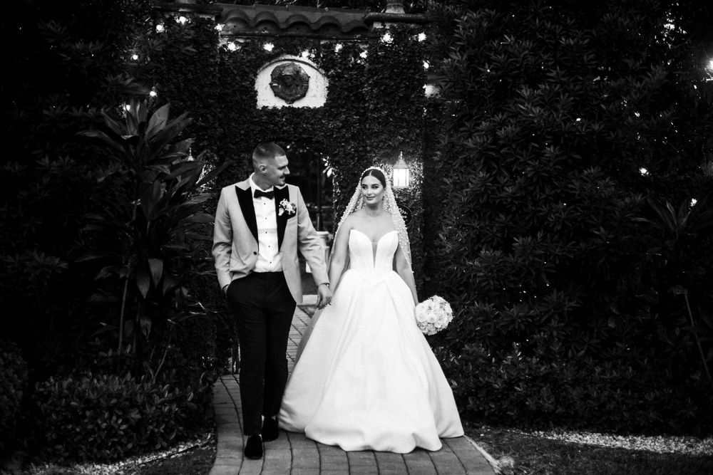 A couple in wedding attire poses for photos in a dark garden setting with twinkling lights in the background.