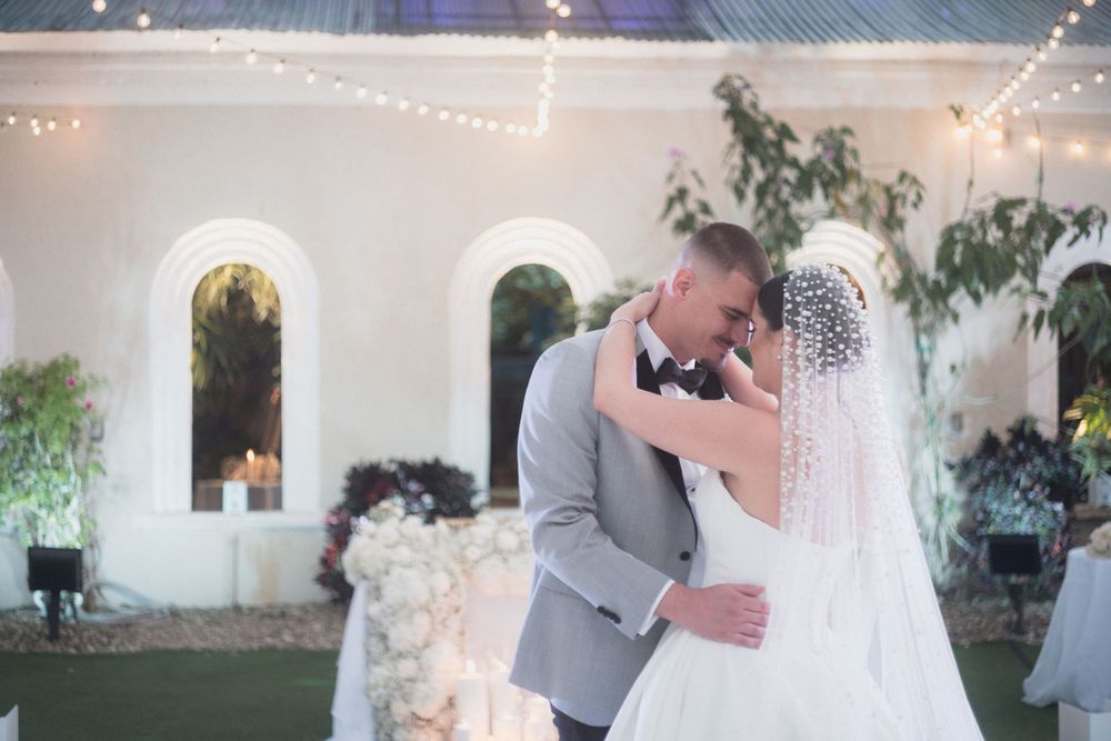 A bride and groom share their first kiss outdoors at a romantic evening wedding reception with string lights and white chapel.