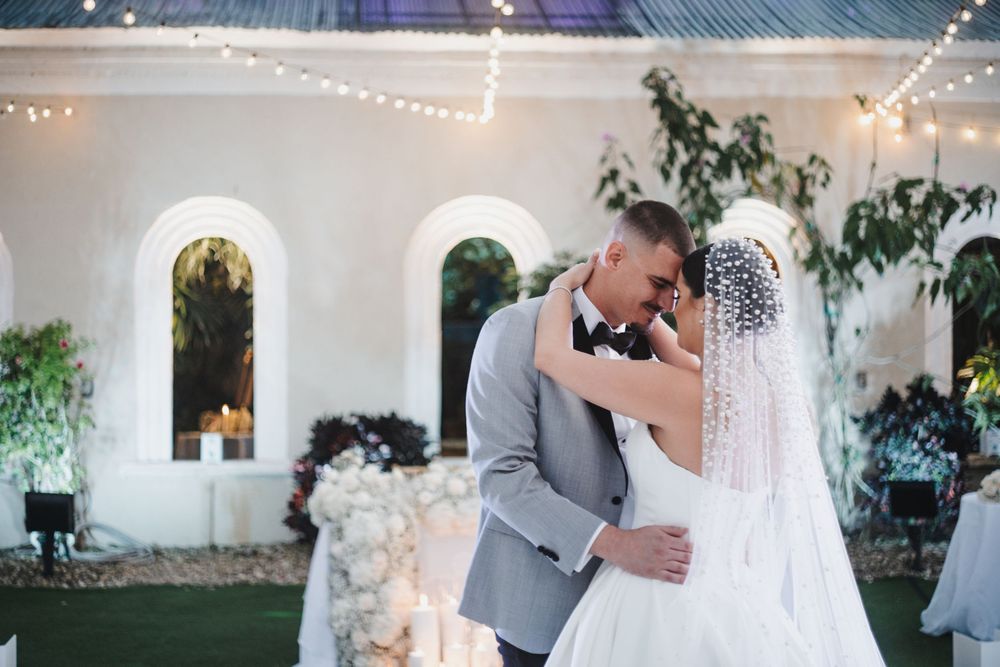 A couple shares their first dance at an elegant white-themed wedding reception with twinkle lights and greenery decor.