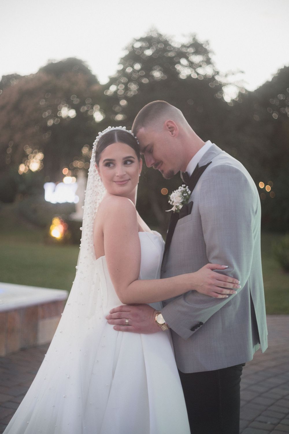 Bride and groom embrace outdoors at dusk, bride wearing white gown with veil, groom in gray suit with boutonniere.
