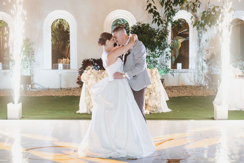 A couple in wedding attire dances together on a white dance floor surrounded by sparkler fountains during their first dance.
