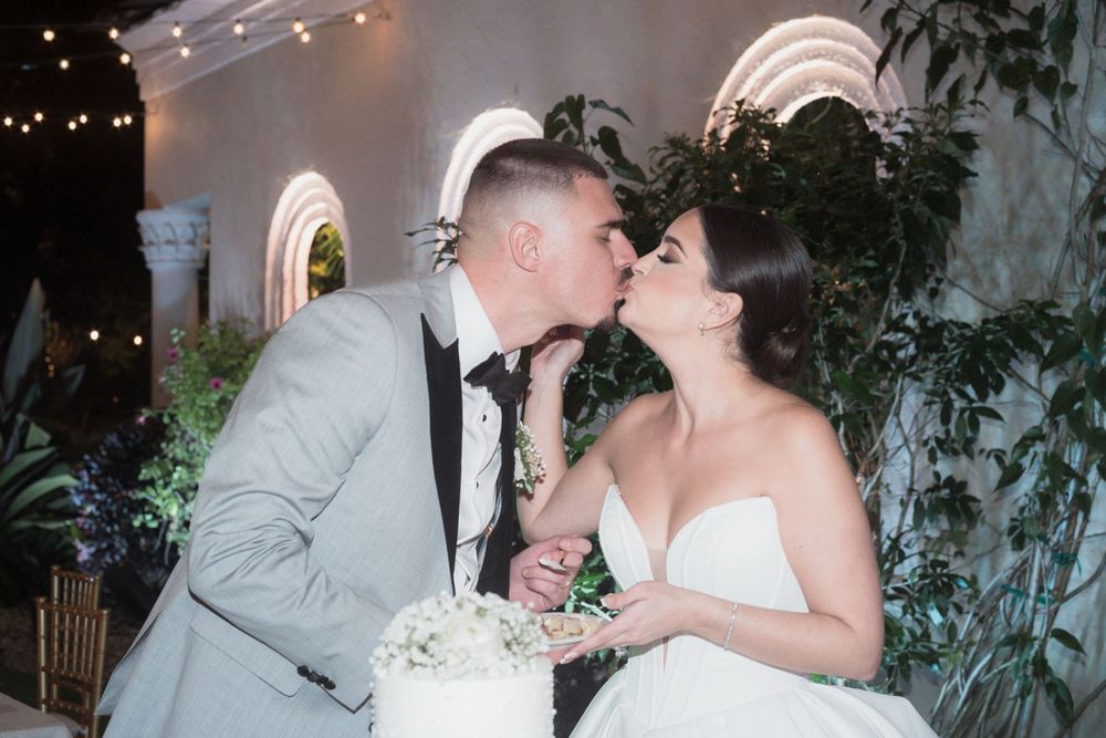 Bride and groom sharing a kiss while cutting their white wedding cake, surrounded by lush greenery and warm string lights.