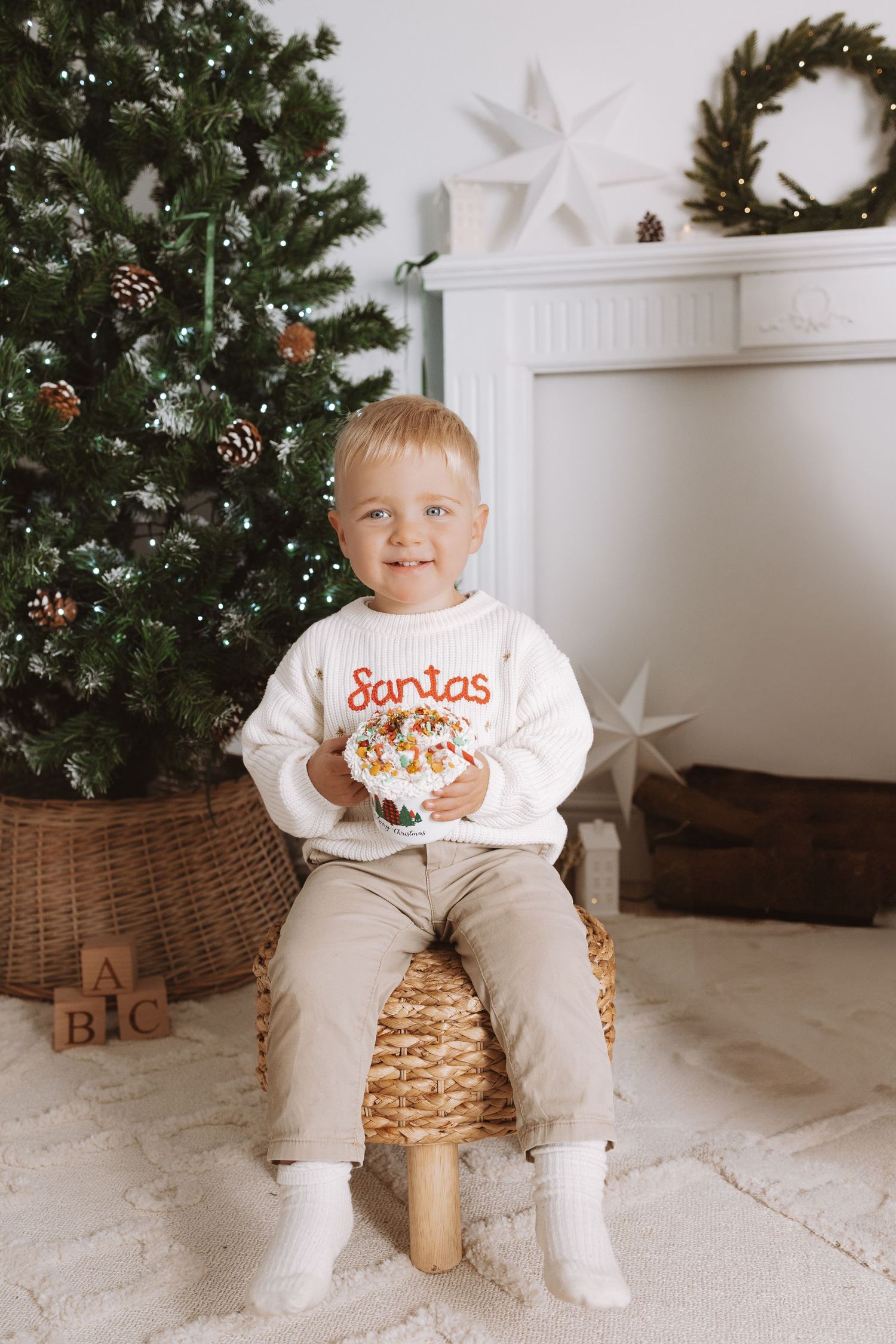 A small child sits on a woven basket in front of a decorated Christmas tree wearing a festive holiday outfit.