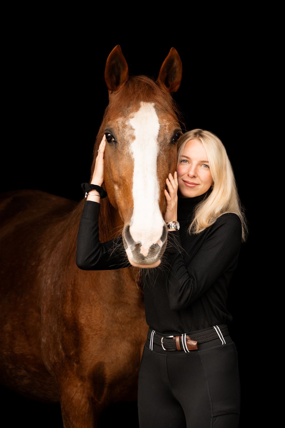 A blonde woman in black equestrian attire affectionately embraces a chestnut horse against a dark background.