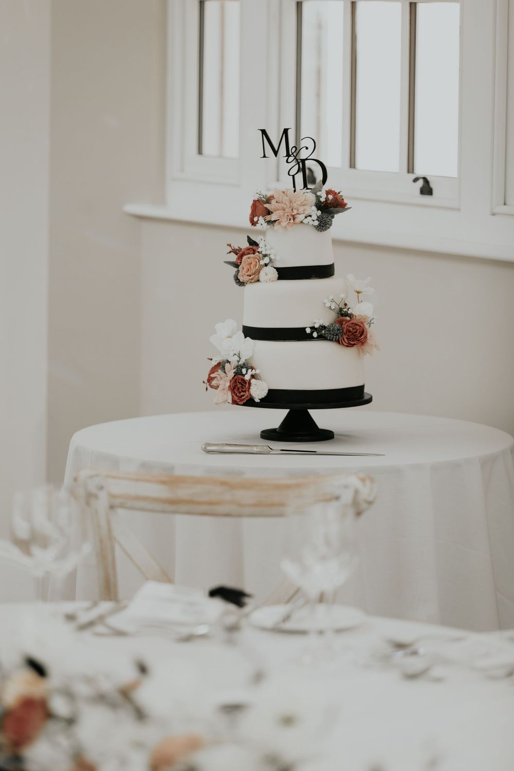 Three-tier white wedding cake with black ribbon, floral accents, and monogram M&D topper on white table.