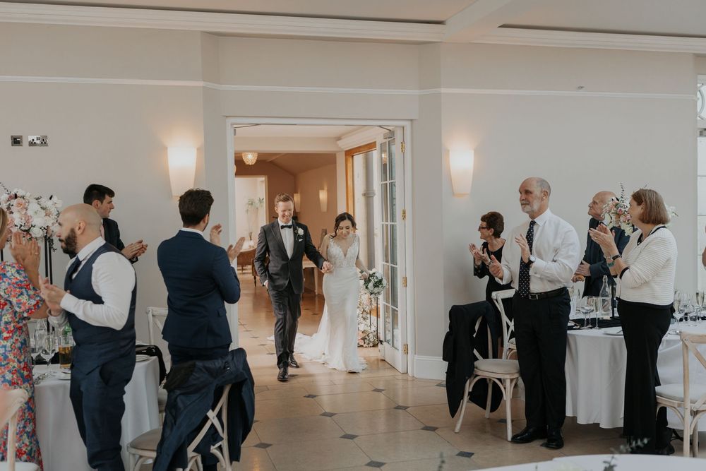 Newlyweds making grand entrance as guests applaud in bright elegant reception venue.