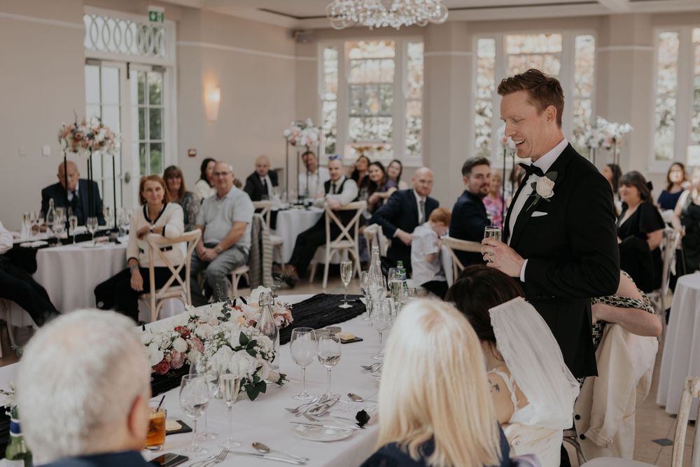 Groom in black tuxedo delivering heartfelt speech to seated wedding reception guests.