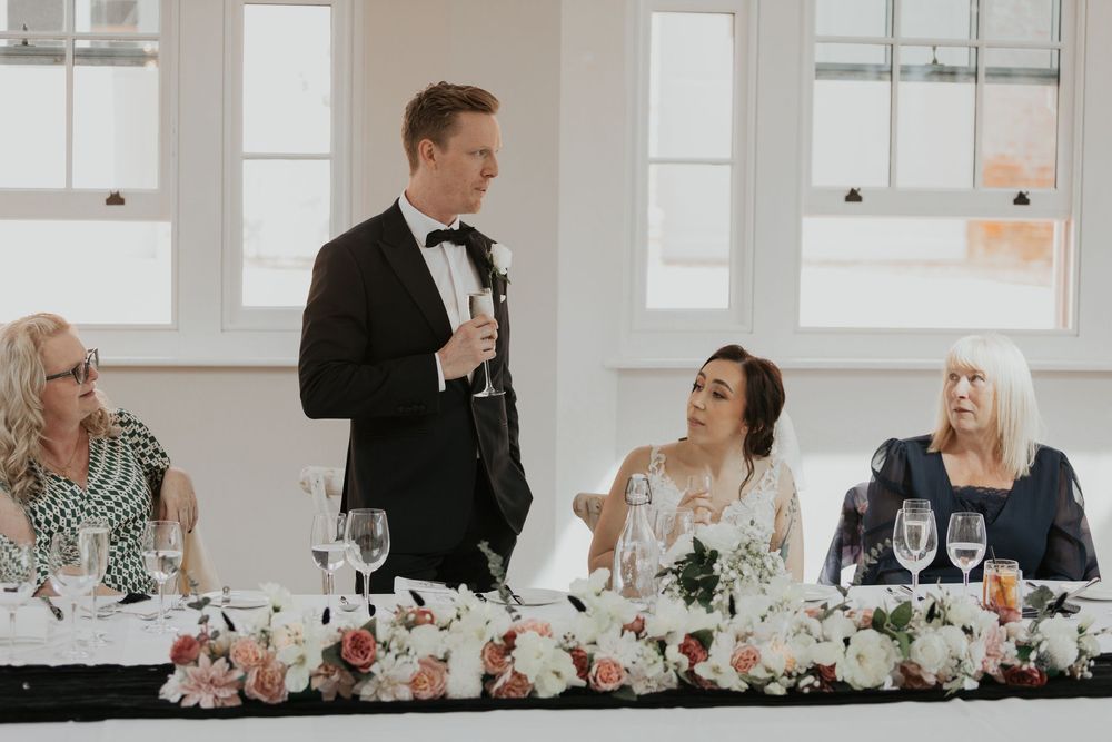 Groom giving wedding speech at floral-decorated head table with bride and family.