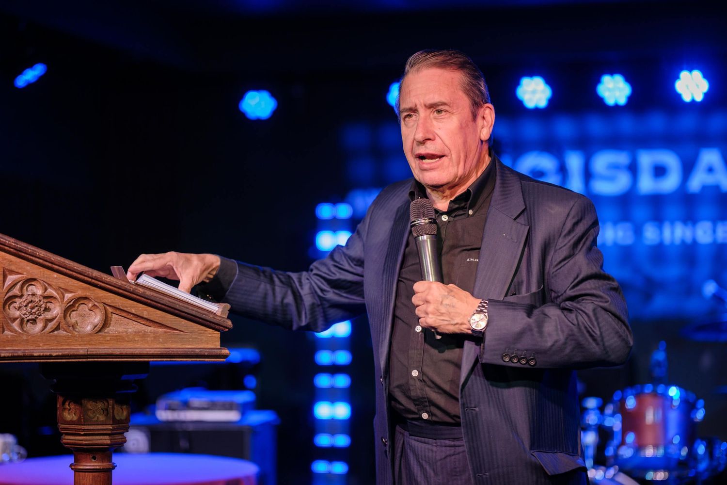A person in a dark suit gestures while speaking on stage with blue lighting and screens visible in the background.